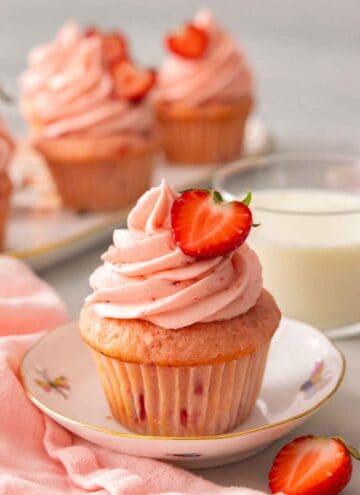 A strawberry cupcake with half a strawberry on top of the pink frosting. More cupcakes and milk in the background.