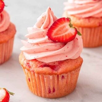 Four strawberry cupcakes with pink strawberry frosting with a cut berry on top.