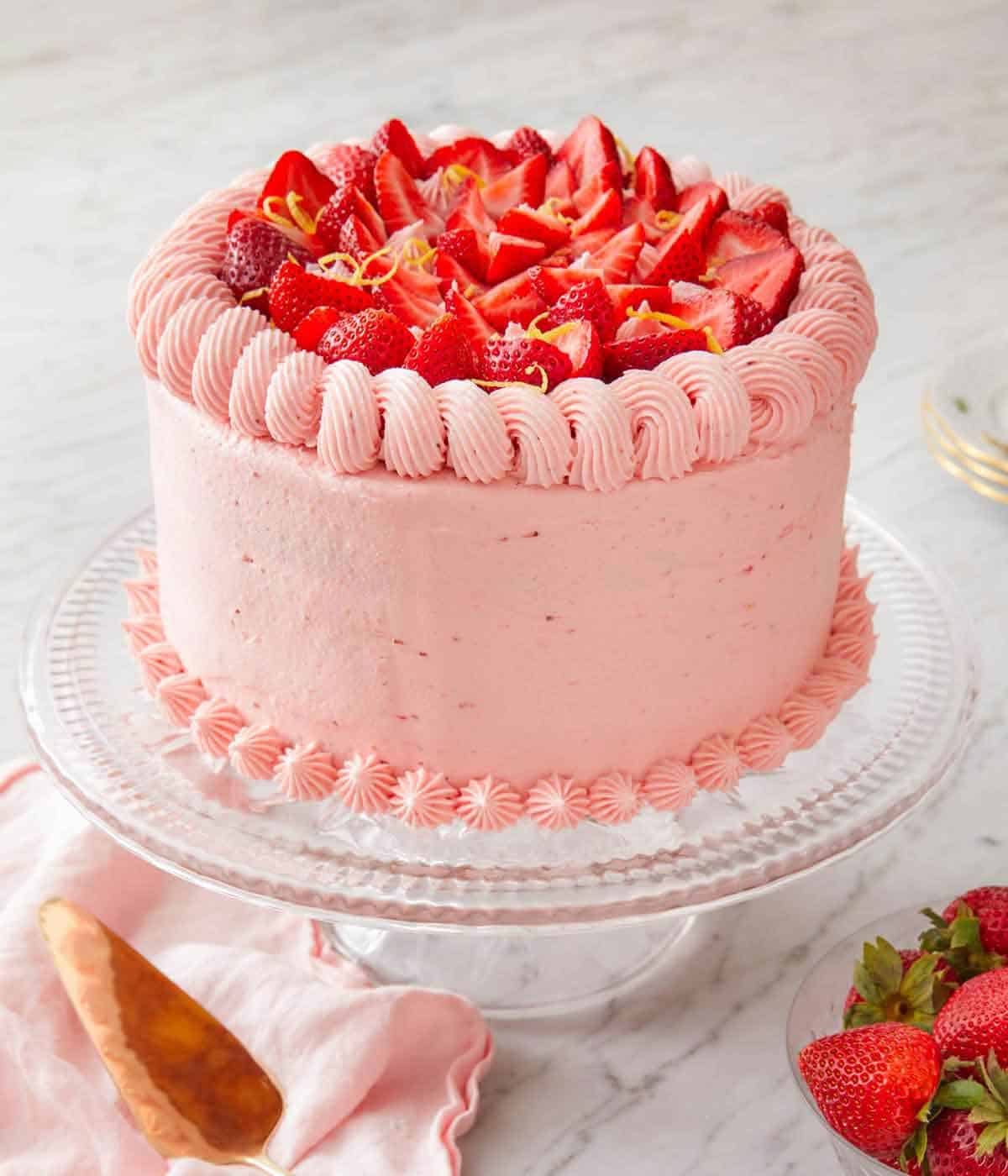 A strawberry lemonade cake on a clear cake stand with some fresh strawberries on top and in a bowl in front.