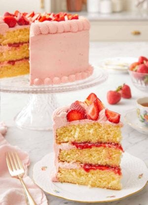 A slice of strawberry lemonade cake on a plate in front of the cut cake in the back on a cake stand.