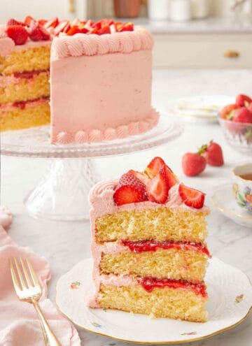 A slice of strawberry lemonade cake on a plate in front of the cut cake in the back on a cake stand.