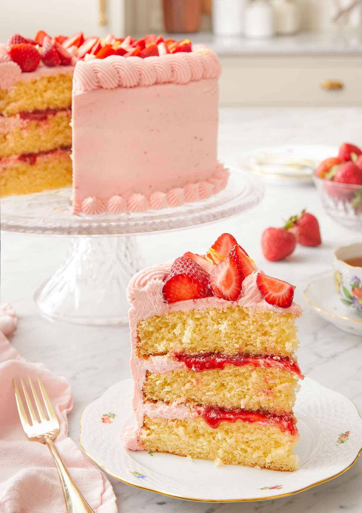A slice of strawberry lemonade cake on a plate in front of the cut cake in the back on a cake stand.