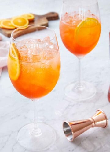 Two stemmed wine glasses of aperol spritzes with a jigger in front and sliced oranges in the background.