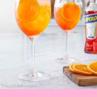Pinterest graphic of two stemmed wine glasses filled with aperol spritz with a bottle of aperol in the background.
