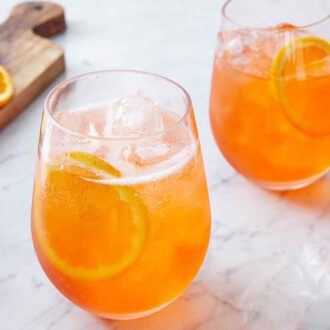Two glasses of aperol spritzes beside a bottle of aperol and sliced oranges.