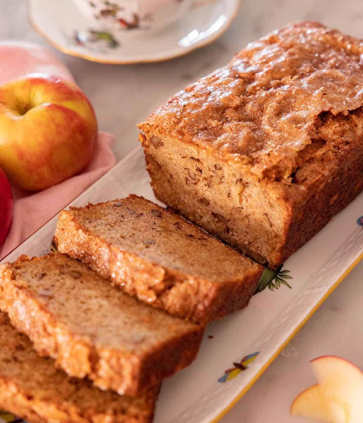 A platter with a loaf of apple bread with three slices cut in front. Apples in the background.