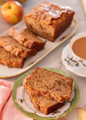 A plate with two slices of apple bread with the rest of the half cut loaf in the background.