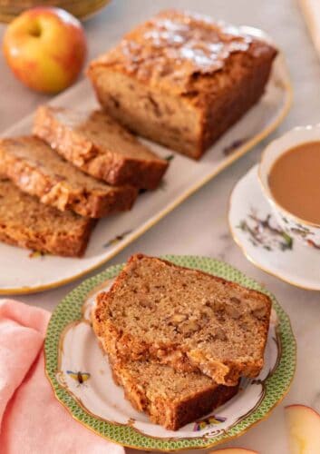 A plate with two slices of apple bread with the rest of the half cut loaf in the background.