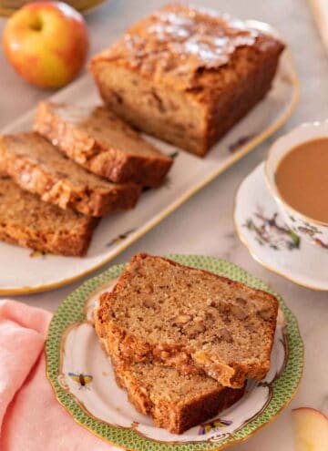 A plate with two slices of apple bread with the rest of the half cut loaf in the background.