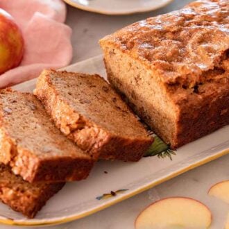A loaf of apple bread on a platter with three slices cut with apples in the background.