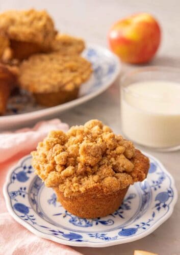 A plate with an apple muffin with a glass of milk in the back with a platter of more muffins in the background.