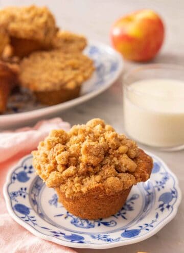A plate with an apple muffin with a glass of milk in the back with a platter of more muffins in the background.