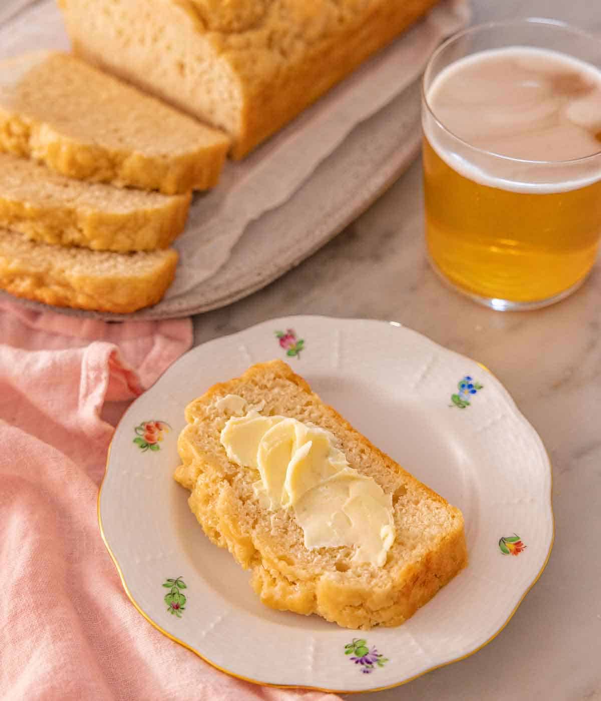 A plate with a slice of beer bread with butter spread on top, with the cut loaf and a drink in the back.