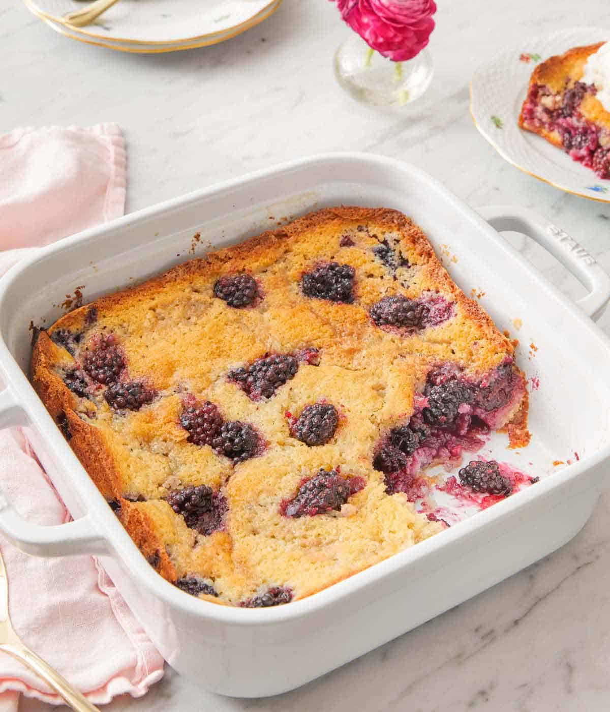 A white baking dish filled with blackberry cobbler with a slice taken out.