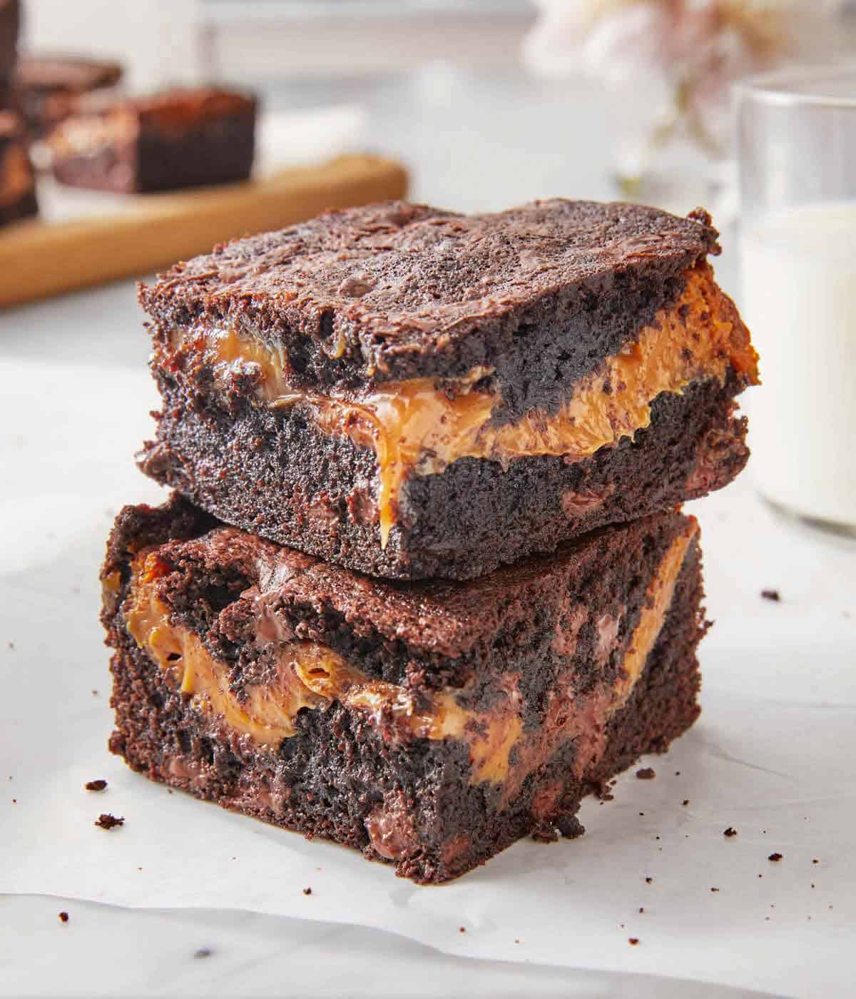 Two caramel brownies stacked on top of each other with a glass of milk in the background.