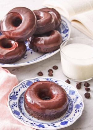 A plate with a chocolate donut by a glass of milk and additional donuts in the back.