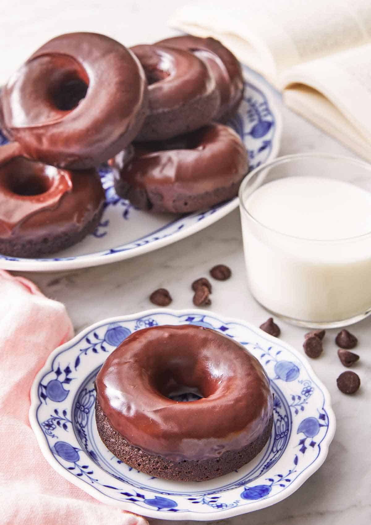 A plate with a chocolate donut by a glass of milk and additional donuts in the back.