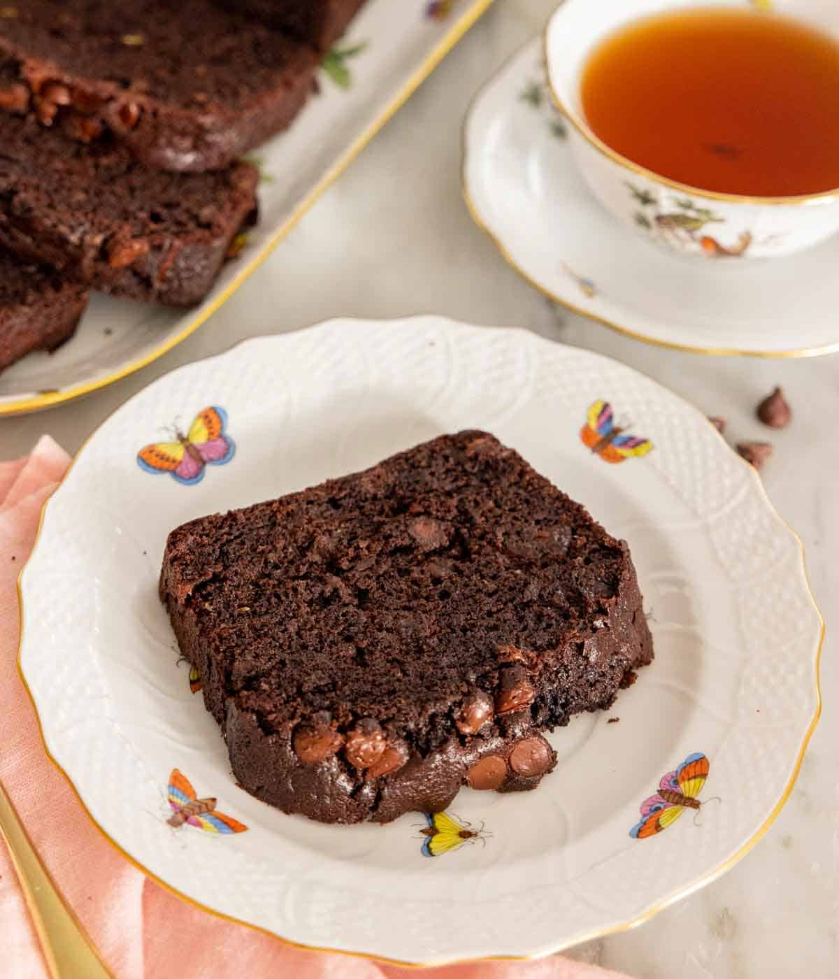 A plate with a slice of chocolate zucchini bread with a cup of tea behind it.