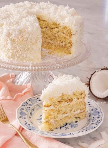 A plate with a slice of coconut cake in front of a cake stand with the rest of the cake.