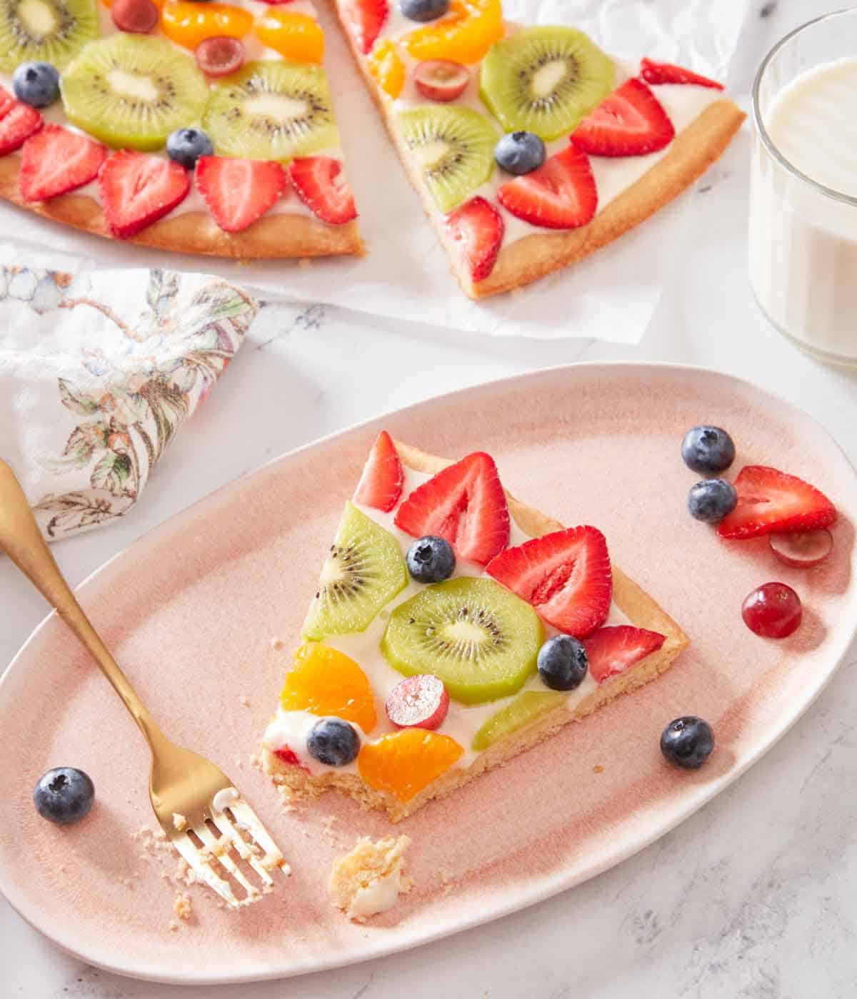 An oval plate with a slice of fruit pizza with a bite taken with more fresh fruit on the plate.