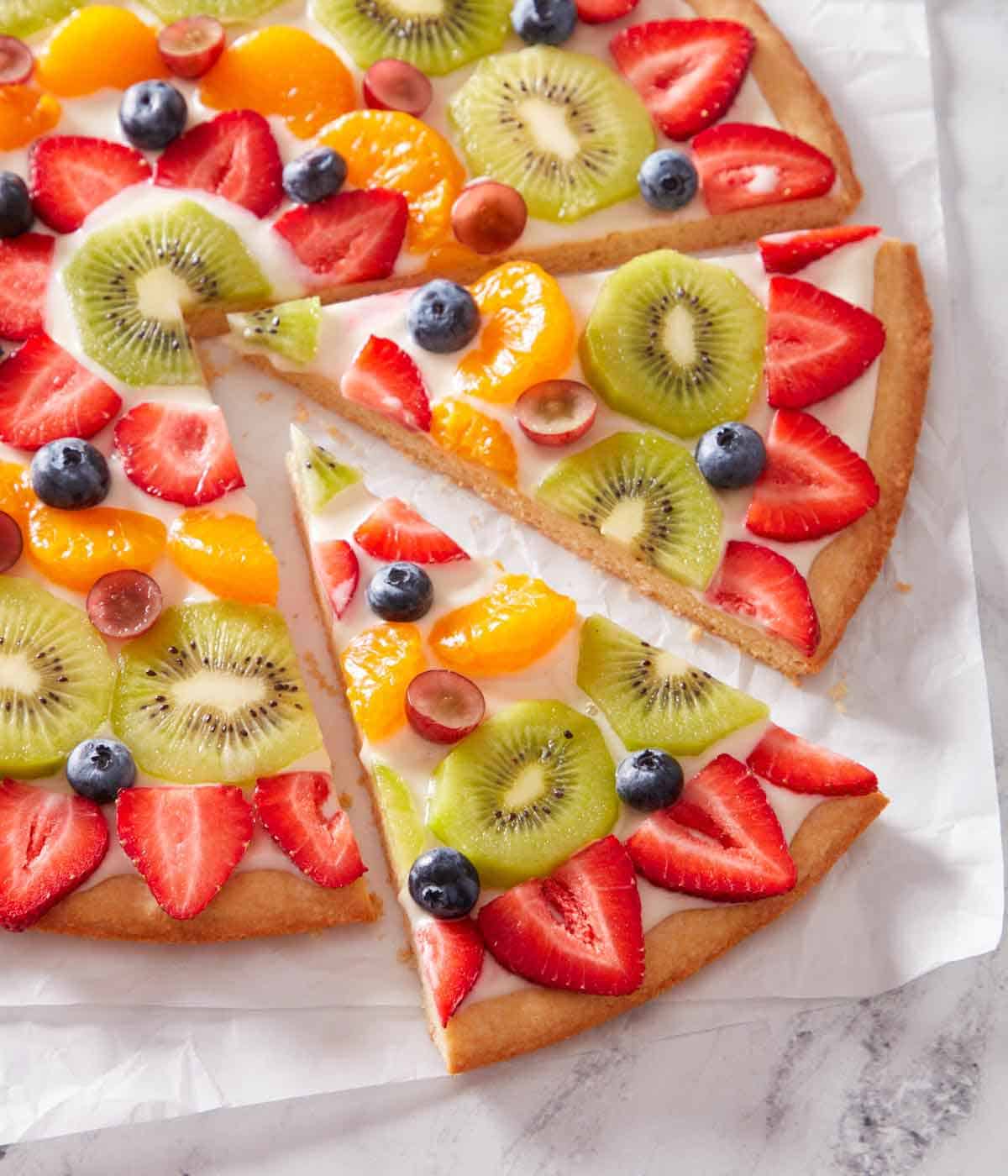 A fruit pizza on parchment paper with two slices cut and slightly pulled out.