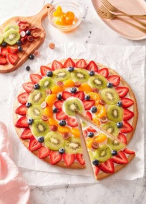 Overhead view of a fruit pizza with a slice cut and pulled forward with more fruit on the side.