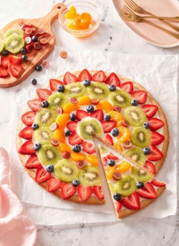Overhead view of a fruit pizza with a slice cut and pulled forward with more fruit on the side.