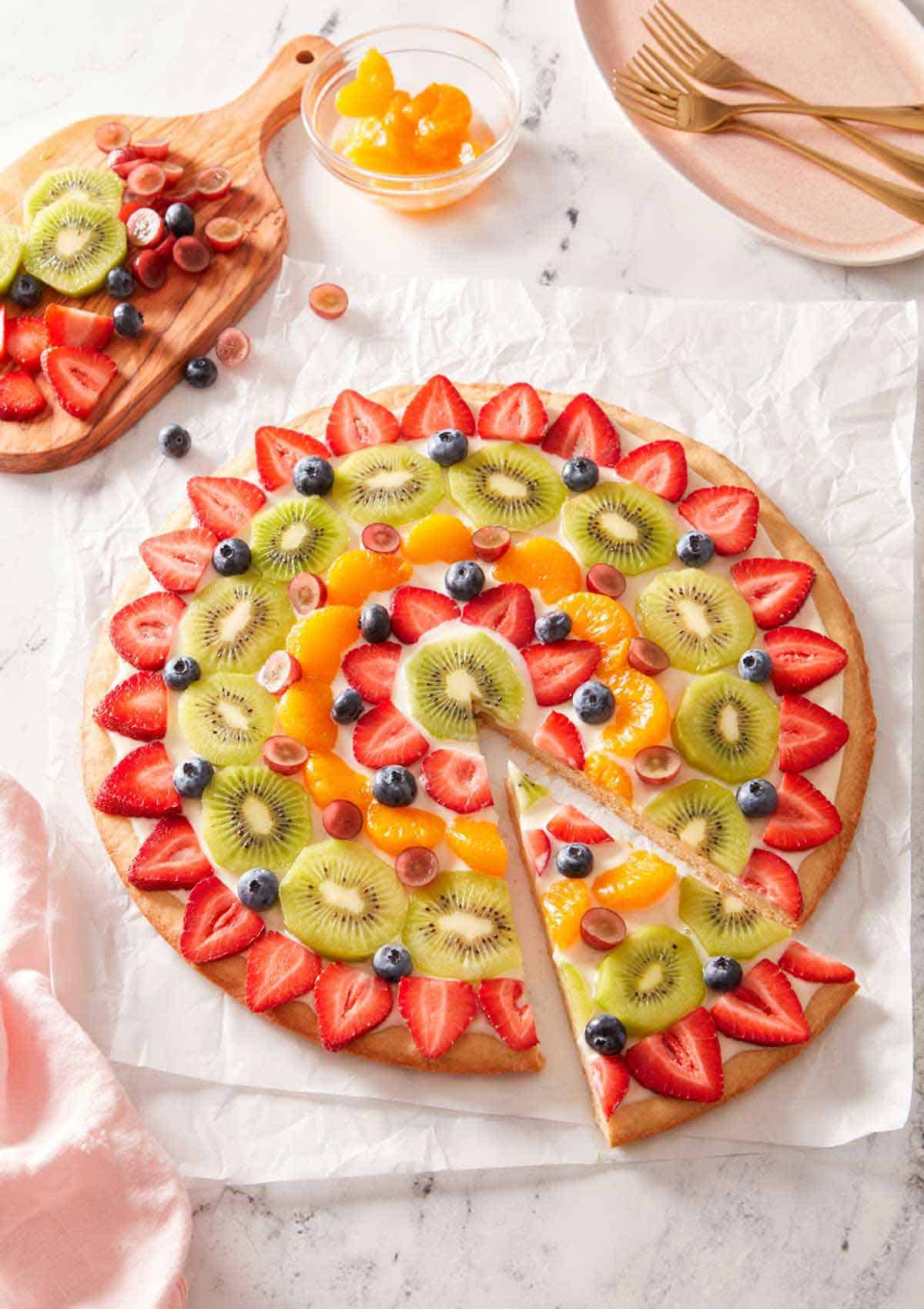 Overhead view of a fruit pizza with a slice cut and pulled forward with more fruit on the side.
