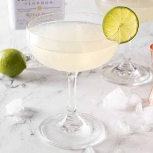 A glass of gimlet with ice, a jigger, and a whole lime behind it.