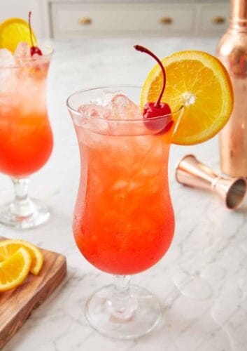 Two hurricane drink with one in the front, both garnished with a cherry and orange slice.