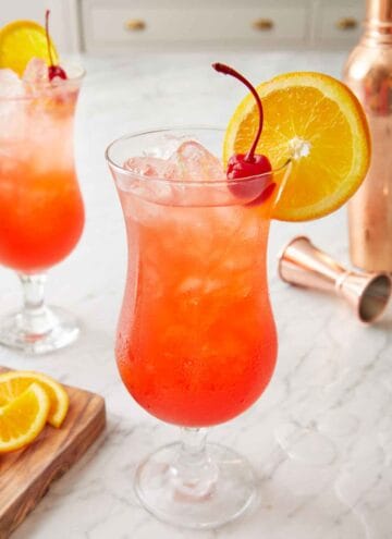 Two hurricane drink with one in the front, both garnished with a cherry and orange slice.