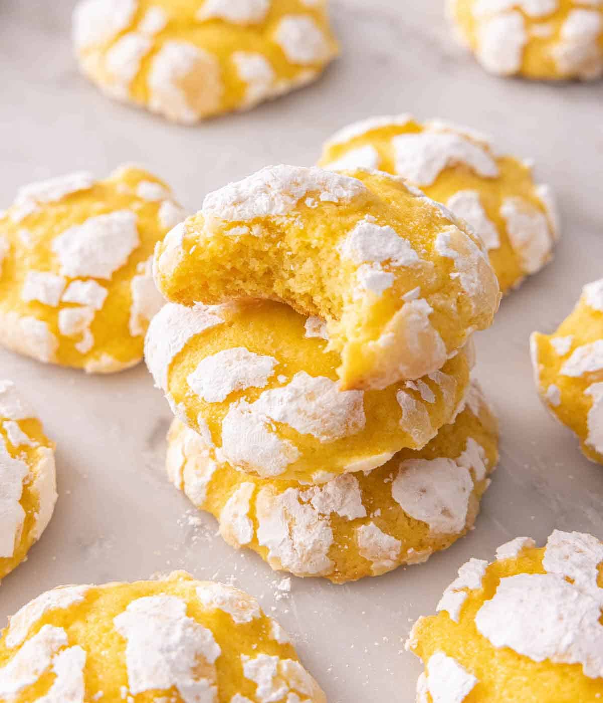 A stack of three lemon crinkle cookies with a bite taken out of the top cookie in the middle of multiple cooks scattered around.