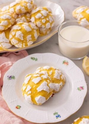 Two lemon crinkle cookies on a plate by a glass of milk in front of a platter of multiple cookies.