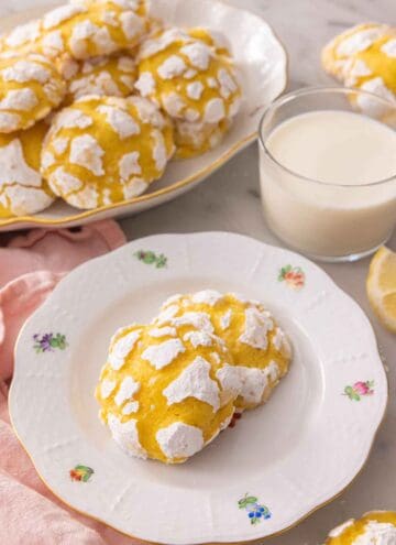 Two lemon crinkle cookies on a plate by a glass of milk in front of a platter of multiple cookies.