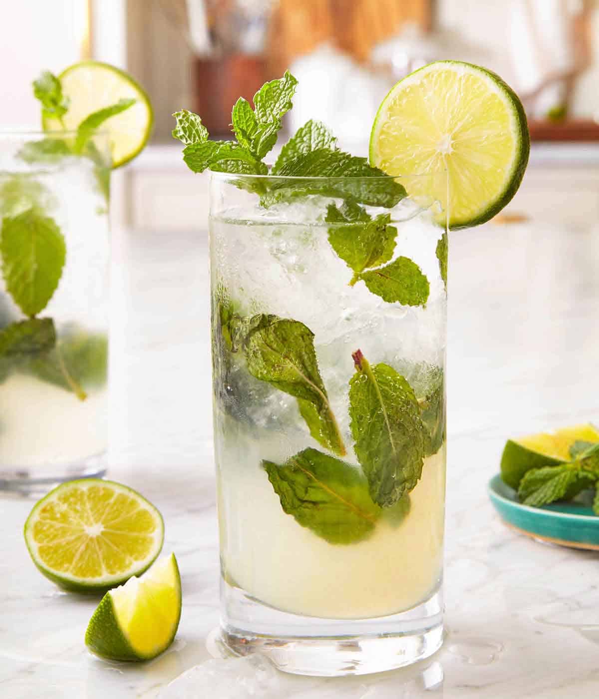 A glass of mojito with fresh lime and mint garnished on the glass and scattered around the counter.