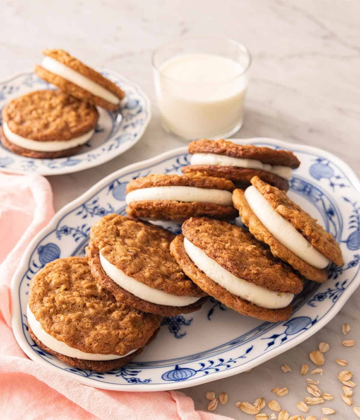 A platter of oatmeal cream pies with two on a plate and glass of milk in the back.