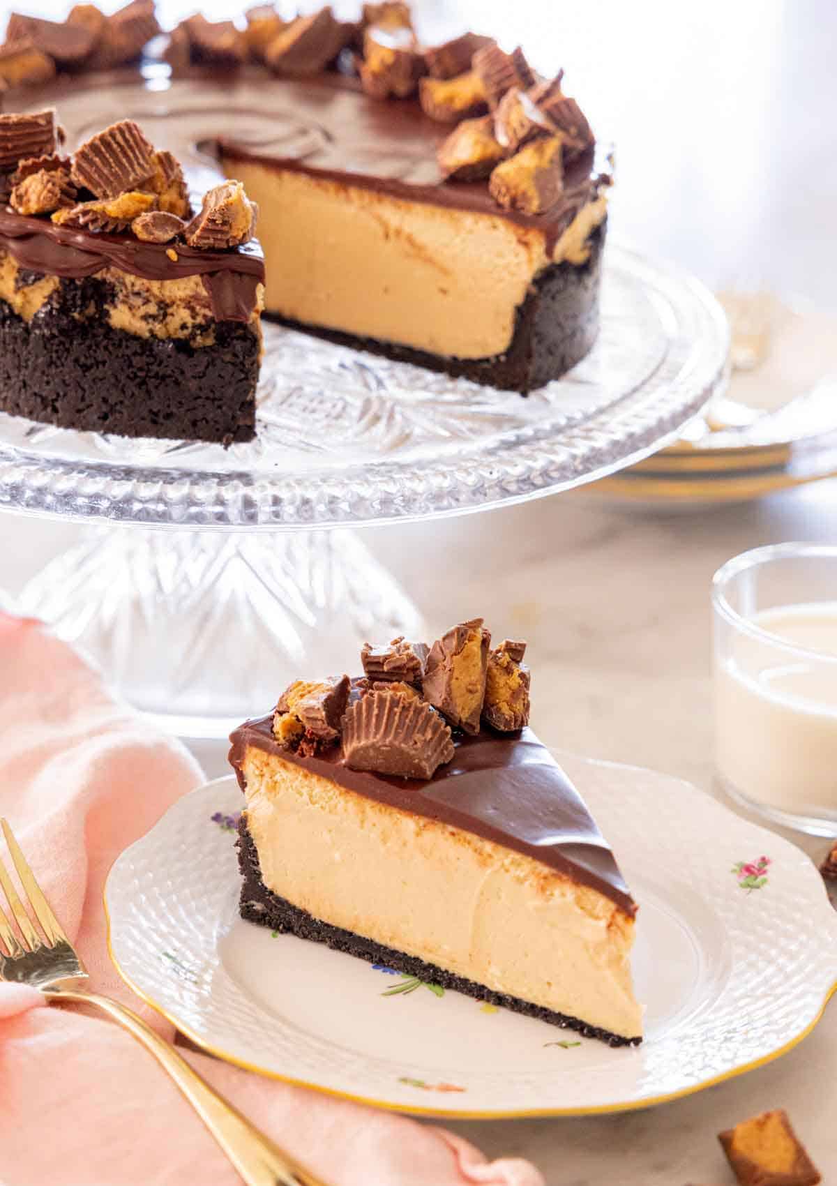 A slice of peanut butter cheesecake on a plate in front of a cake stand with the rest of the cake.