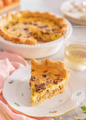 A plate with a slice of quiche Lorraine with the rest of the quiche in the background.
