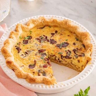 A pie dish with a quiche Lorraine with a slice cut out, plated in the backgrounf.