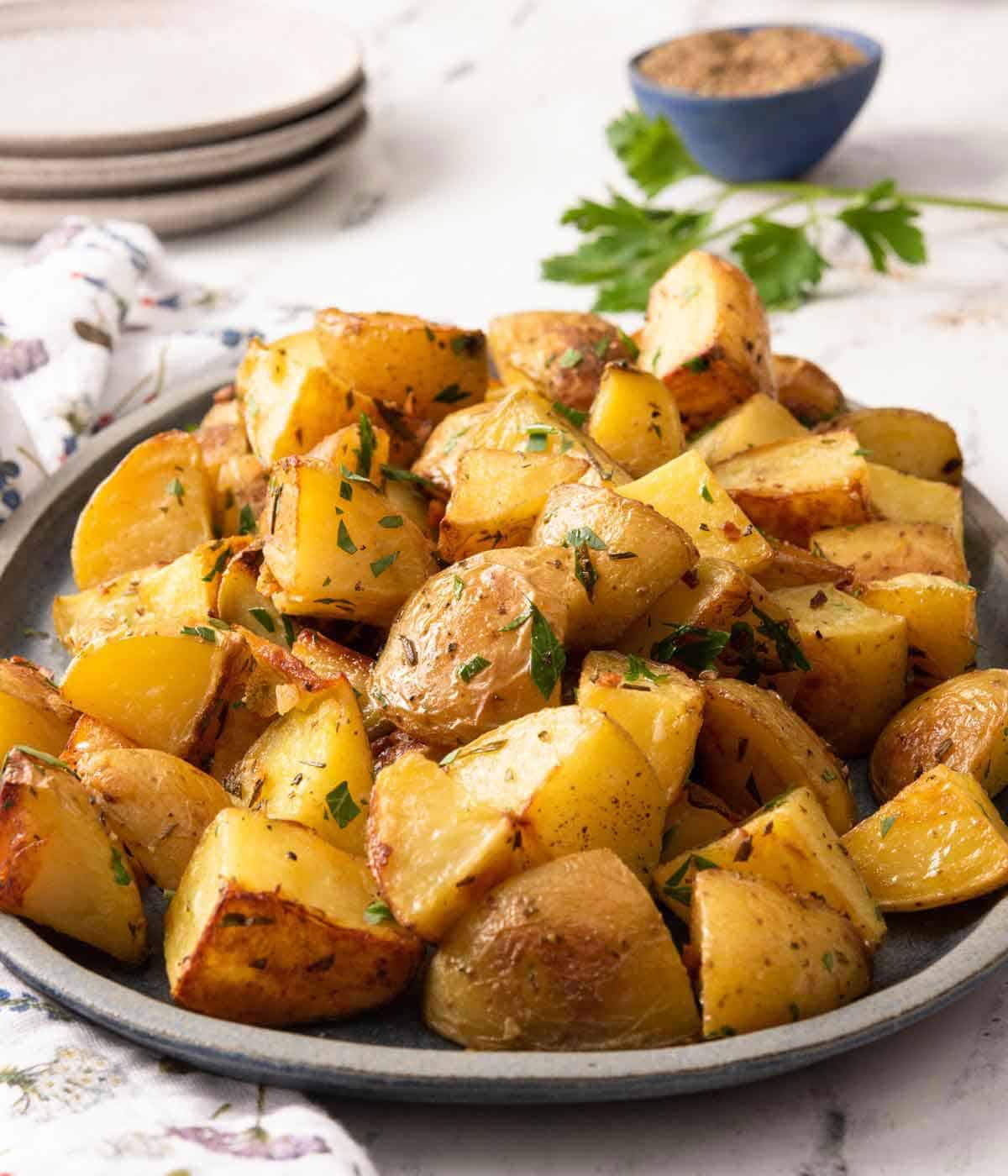 A platter of roasted potatoes with chopped parsley as garnish and a pinch bowl of pepper in the back.