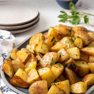Pinterest graphic of a platter of roasted potatoes with a stack of plates, small bowl of pepper, and fresh parsley in the background.