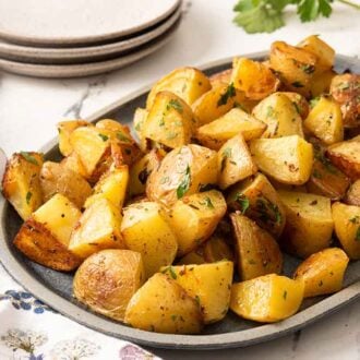An oval platter of roasted potatoes with fresh chopped parsley on top with a stack of plates in the background.
