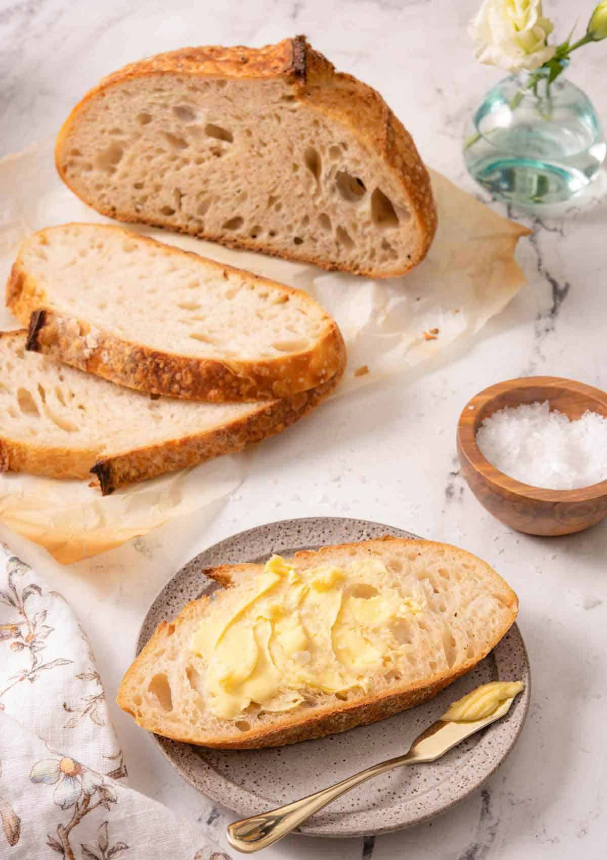 A plate with a slice of buttered sourdough bread with a loaf in the back with two slices cut. Pinch bowl of salt on the side.