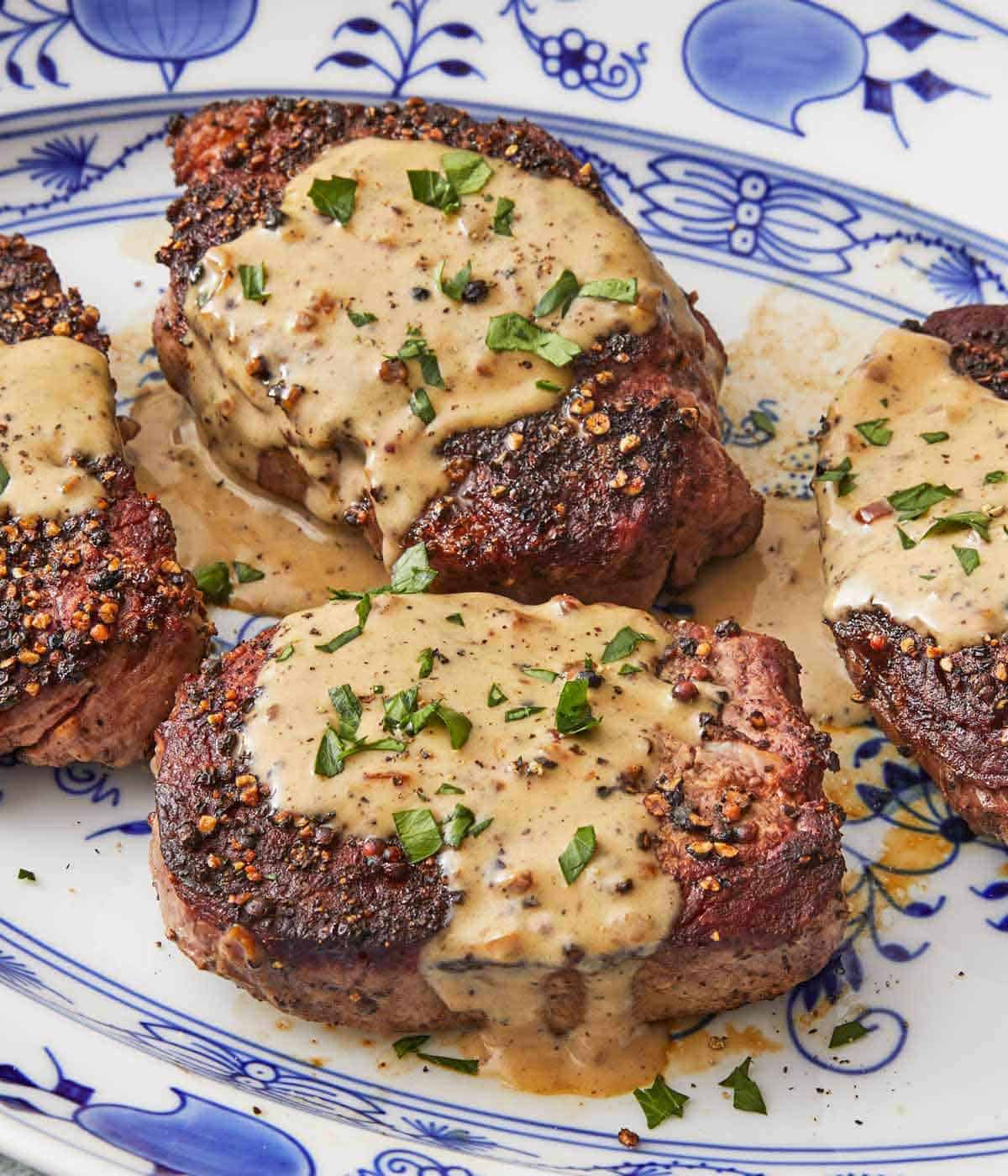 Close up of multiple steak au poivre on a serving plate with herb chopped herbs on top.