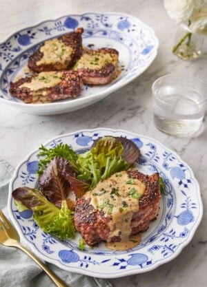 A plate of steak au poivre with a side of salad in front of a platter with three steak au poivre.