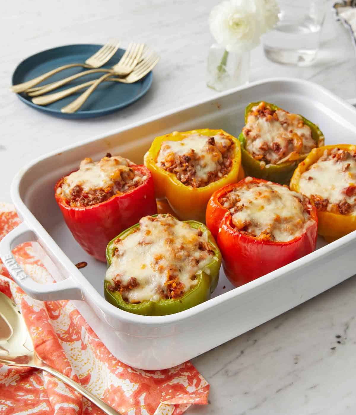 A white baking dish with six stuffed peppers with a plate with multiple forks in the background.