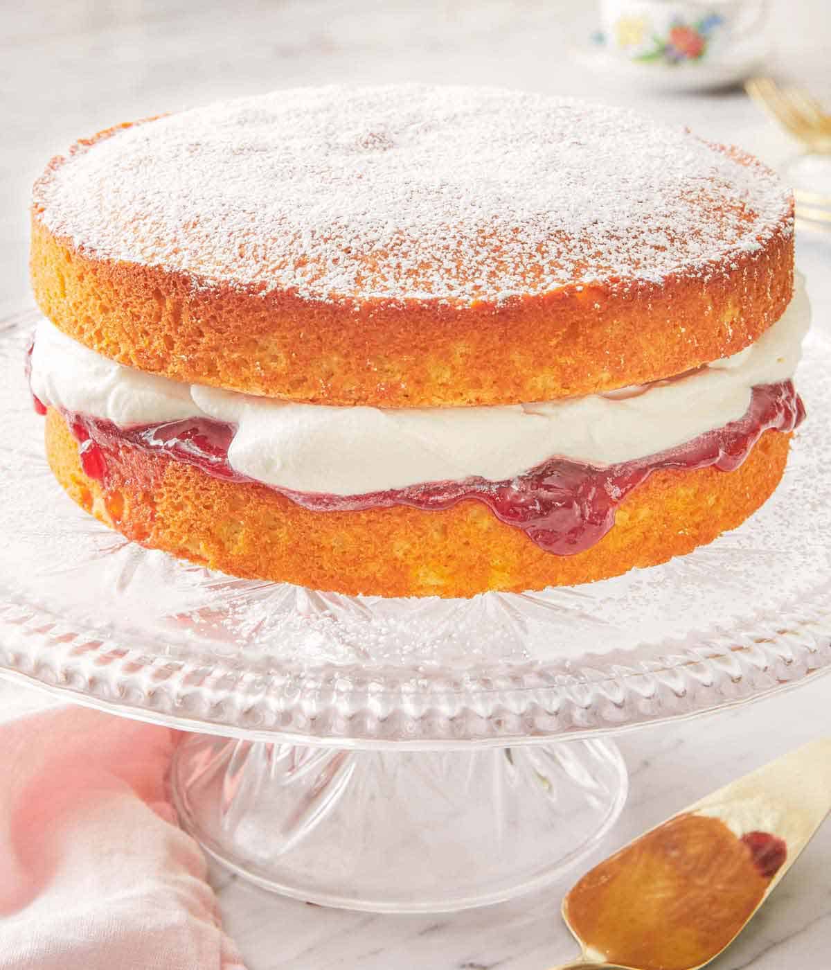 A Victoria sponge cake on a clear cake stand with the jam and whipped cream dripping out.