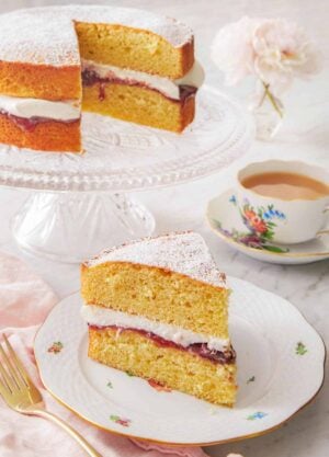 A plate with a slice of Victoria sponge cake in front of a cup of coffee and the rest of the cake.