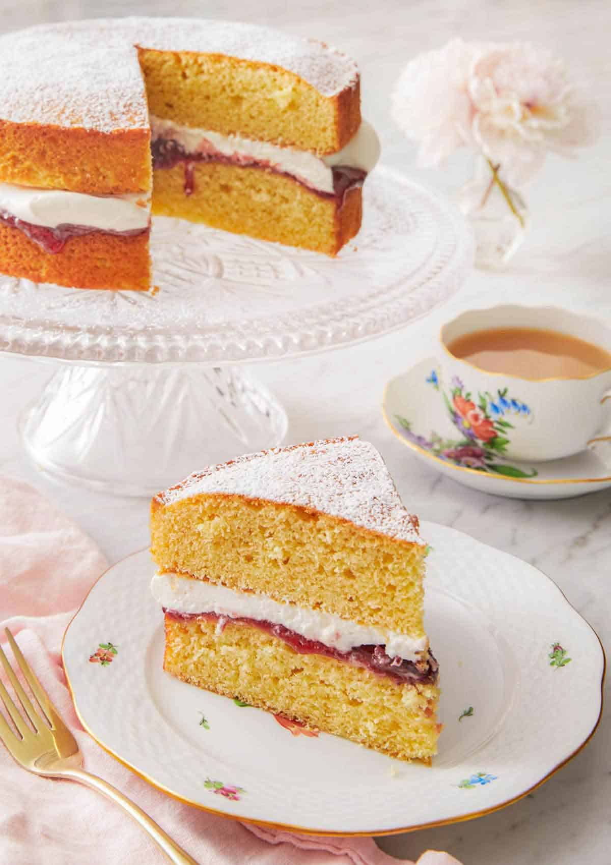 A plate with a slice of Victoria sponge cake in front of a cup of coffee and the rest of the cake.