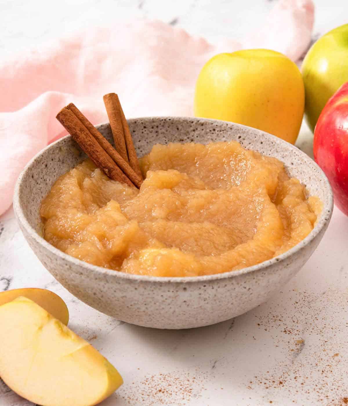 A bowl of applesauce with two cinnamon sticks stuck inside with apples around it.